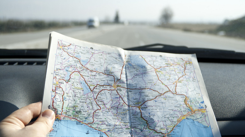 A hand holds up a paper map in front of a dashboard of a car looking out onto a highway.