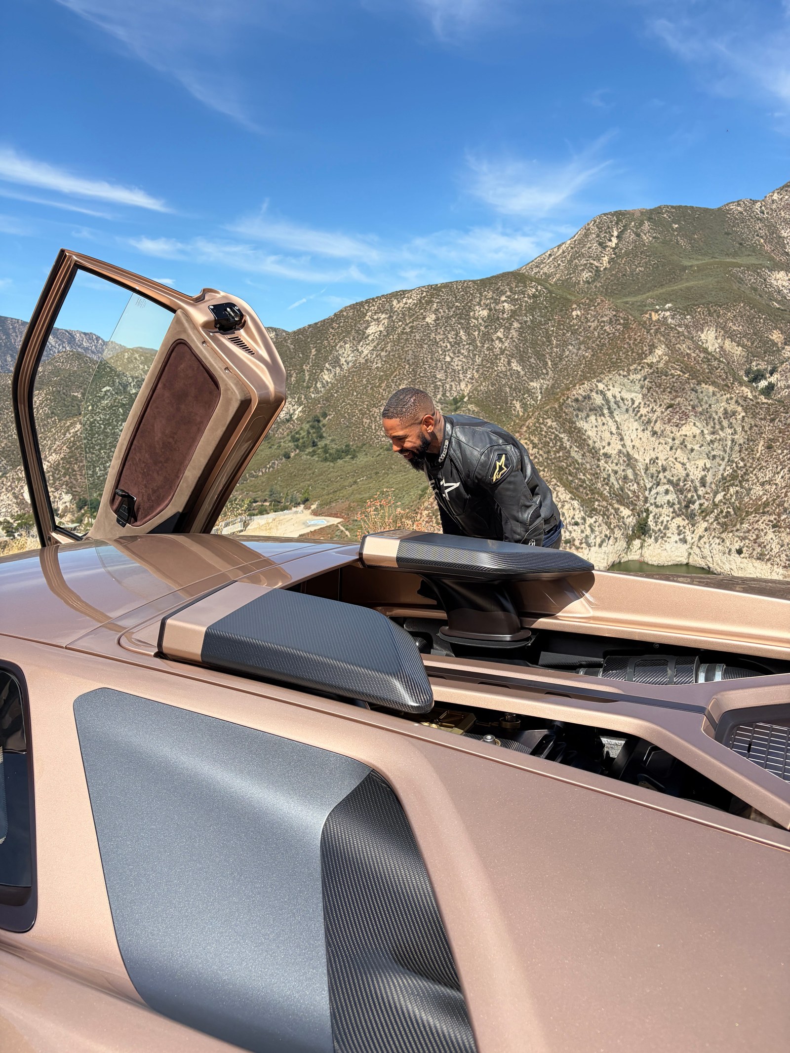 Man in a leather jacket leaning over the open roof of a sleek car with mountainous scenery in the background. Man in a leather jacket leaning over the open roof of a sleek car with mountainous scenery in the background.