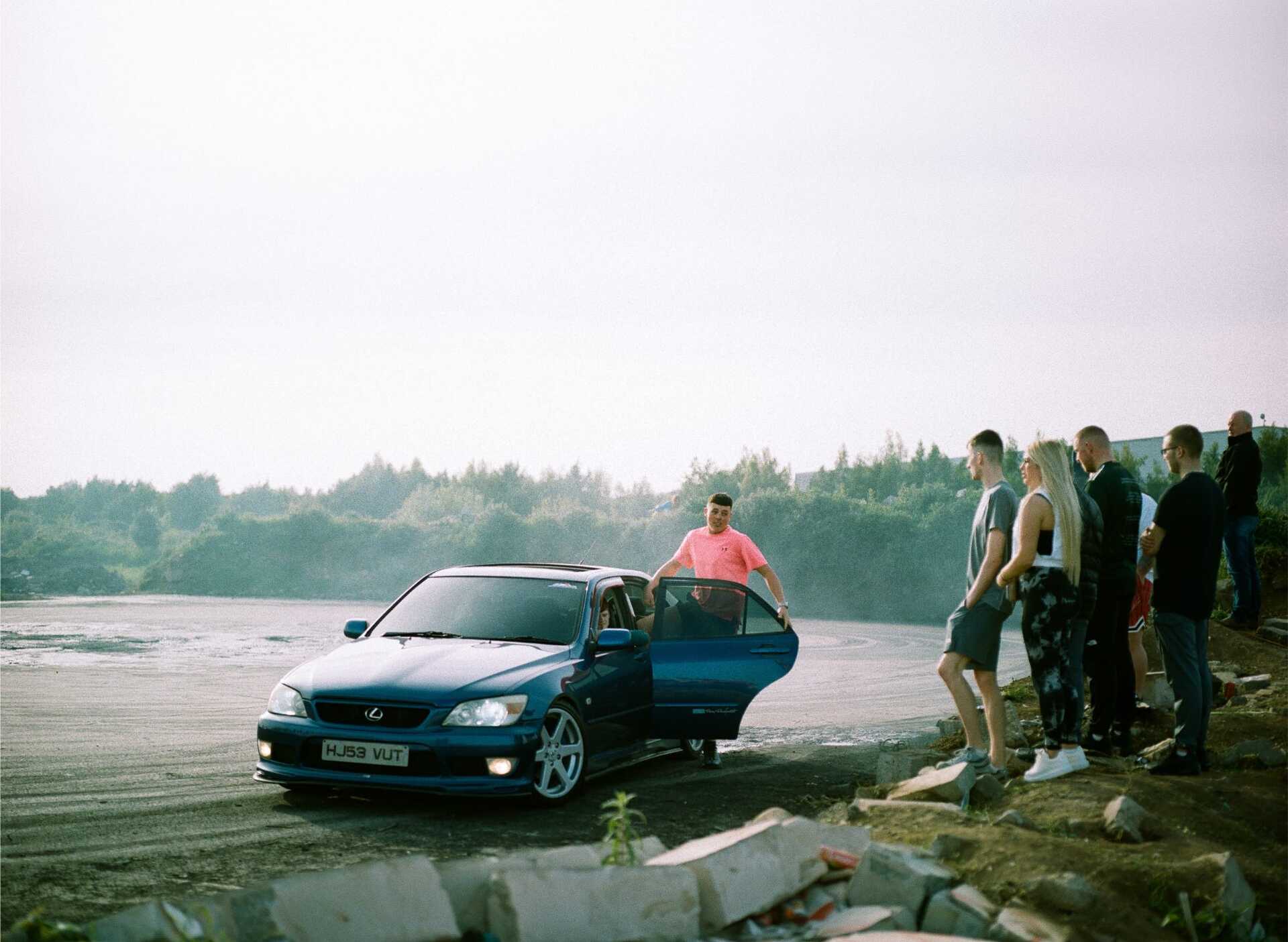 A group of people watching a blue car near an empty lot with trees in the background and smoke in the air.