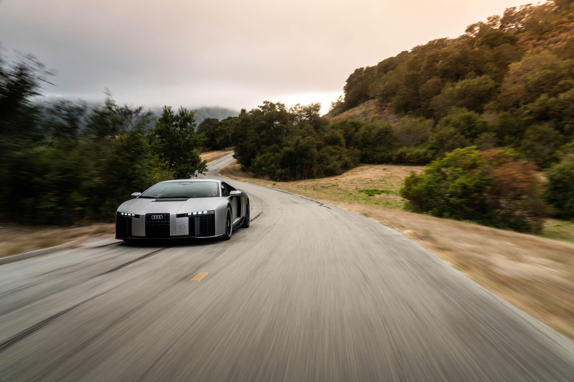 A sleek gray car navigating a winding road surrounded by greenery and hills.