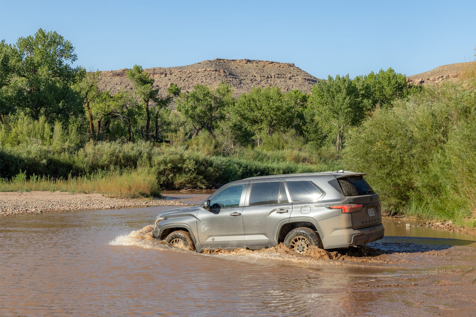A gray SUV driving through a muddy river surrounded by greenery and hills.