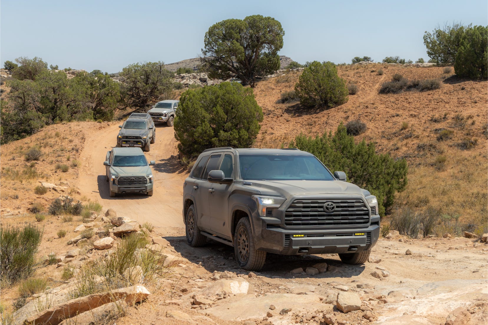 A convoy of off-road vehicles navigating a rugged dirt trail surrounded by desert terrain and sparse vegetation.