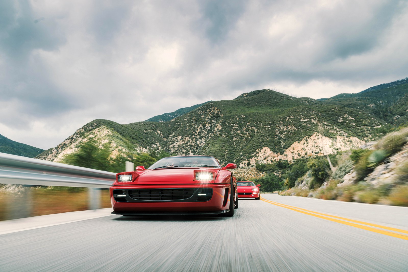 Two red sports cars driving on a winding road surrounded by mountains under a cloudy sky.