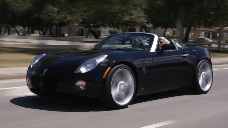 Front 3/4 view of Jay Leno driving a black Pontiac Solstice in front of trees and a neighborhood