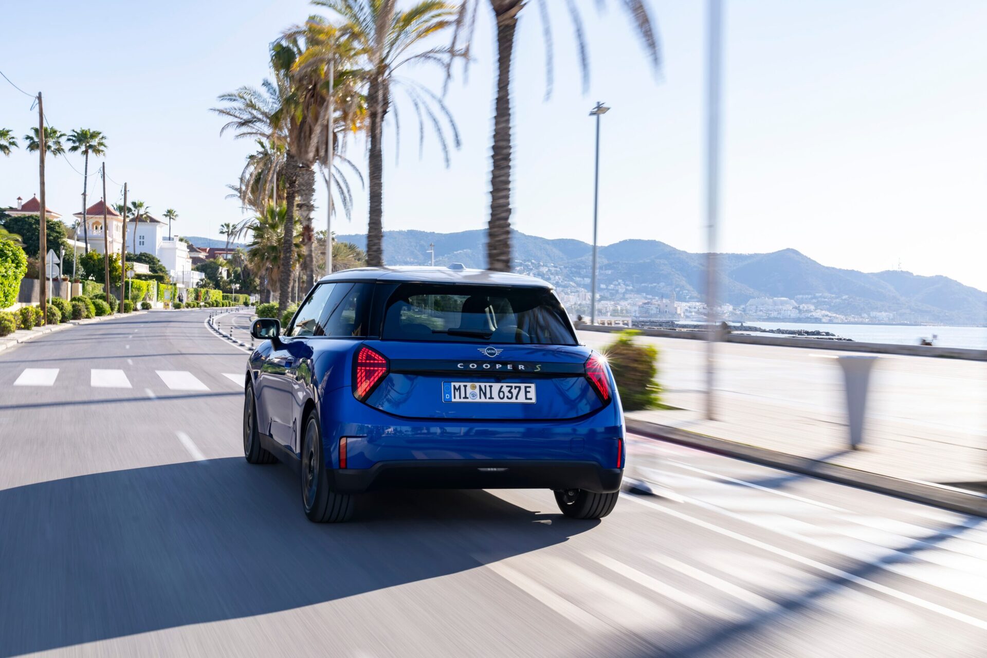 A blue car driving along a palm tree-lined coastal road with mountains in the background.