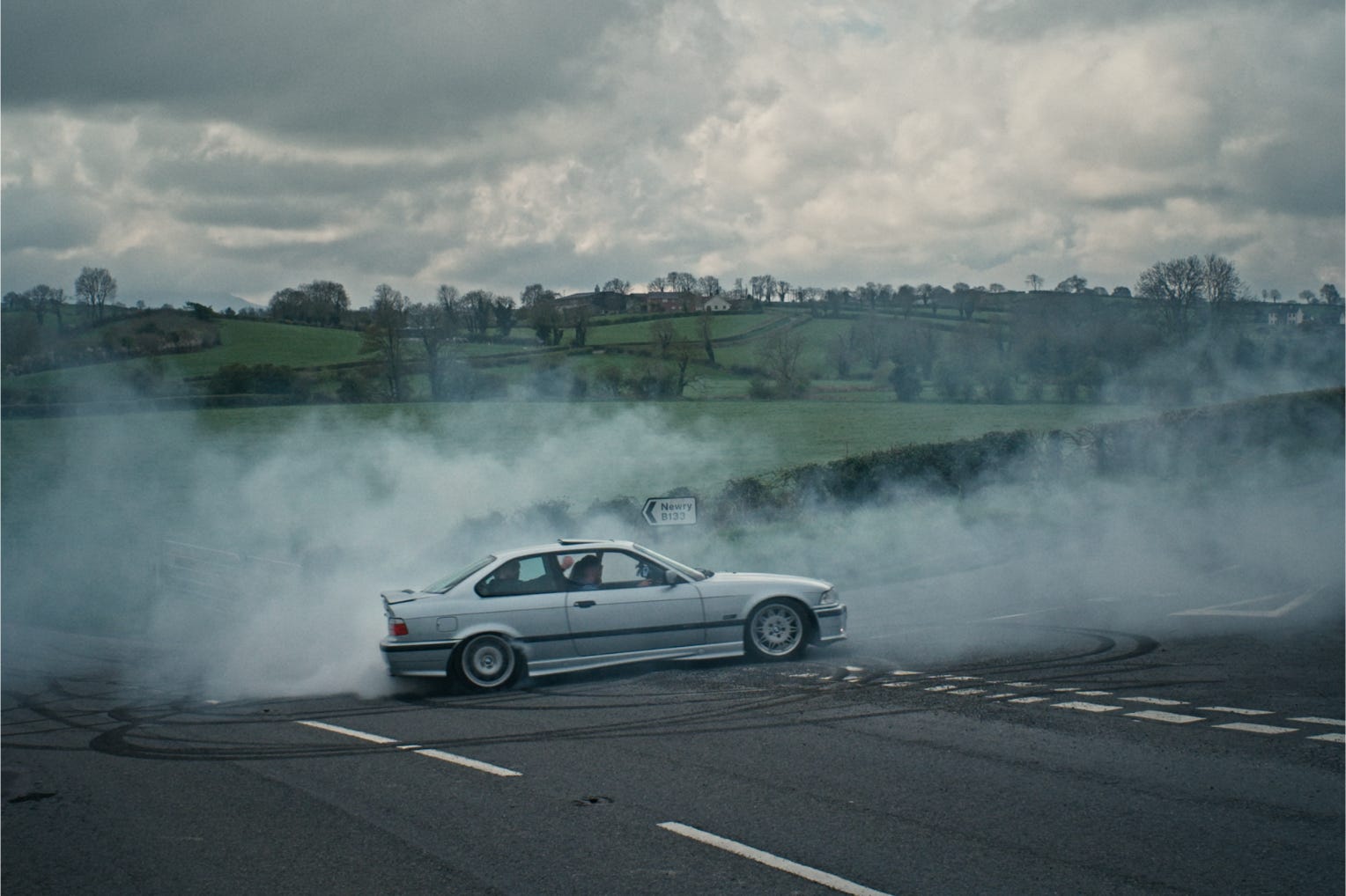A gray car performing a burnout on a rural road with smoke and green fields in the background.