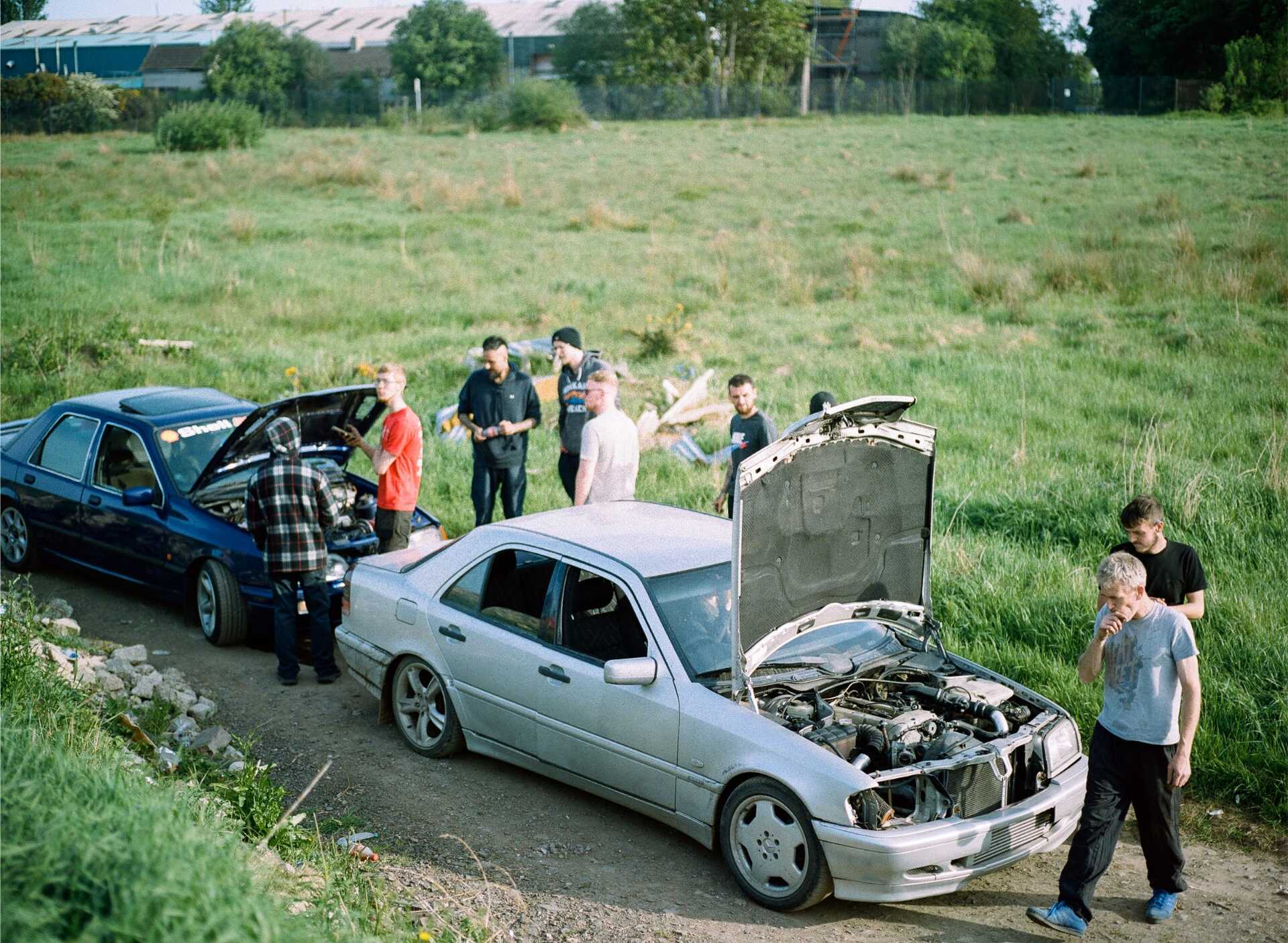 Group of people gathered around two cars with hoods up, inspecting engines in a grassy area.