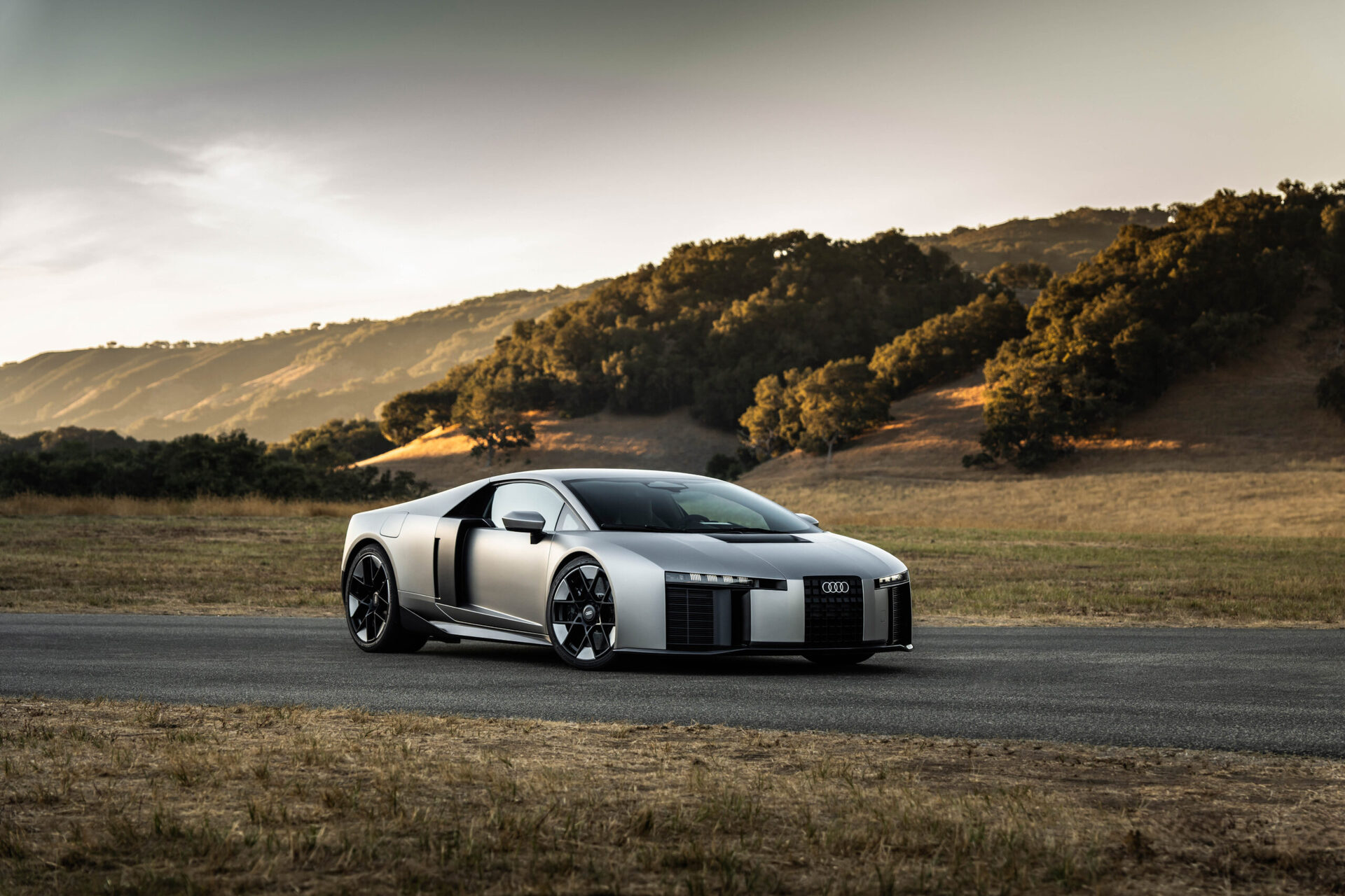 Sleek silver sports car parked on a road with a scenic landscape of hills and trees in the background.