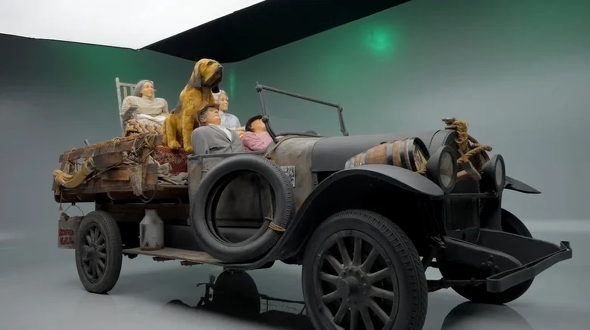 Front-three-quarter view of the rustic Beverly Hillbillies truck prop on a green screen studio set, loaded with family mannequins, tied-down household goods, and a large hound dog statue.