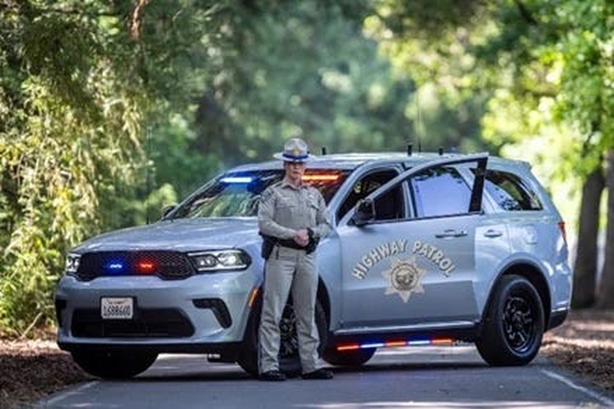 highway patrol officer standing next to patrol suv with open door on a tree lined road.