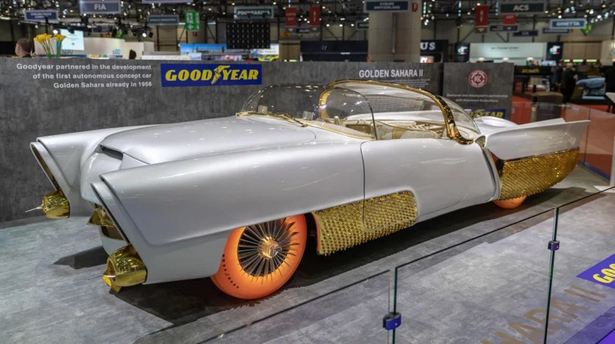 Front-three-quarter view of the Golden Sahara II custom concept car on an exhibit stage, featuring a pearl white body, gold-textured side skirts, a twin-bubble canopy, and illuminated orange translucent Goodyear tires.