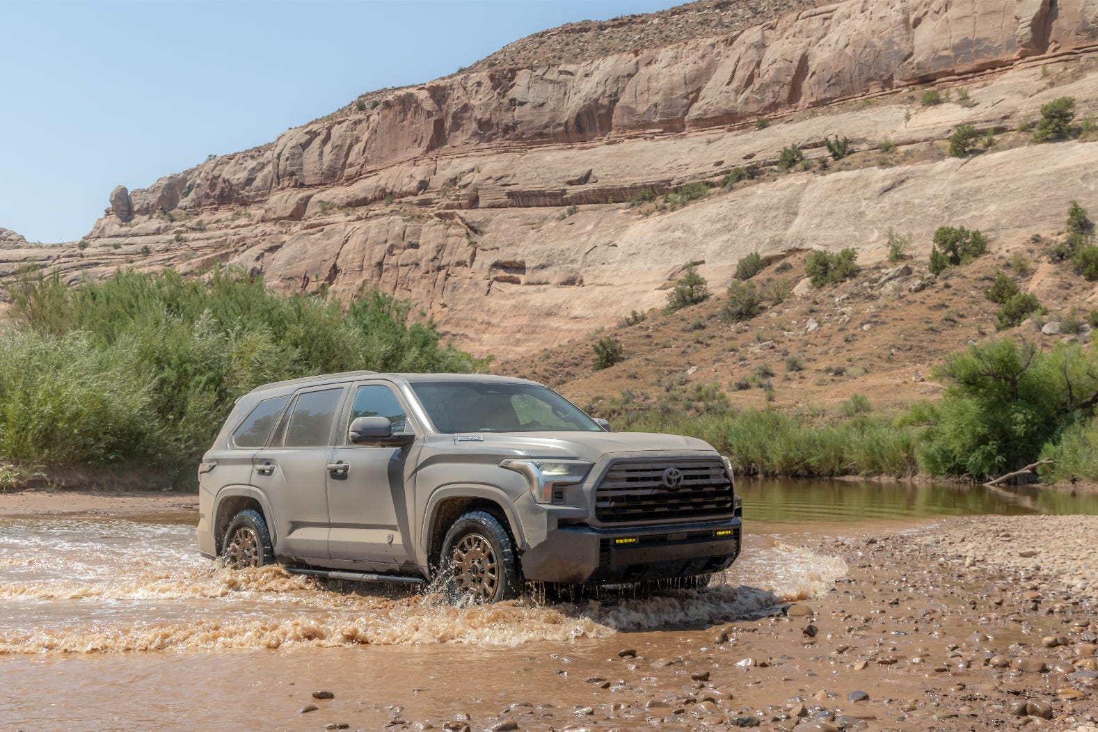 Off-road vehicle navigating through shallow water with rocky terrain and rugged cliffs in the background.