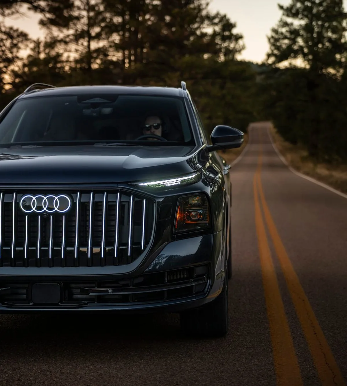 Black SUV on a scenic road surrounded by trees at sunset, with a driver wearing sunglasses visible through the windshield.